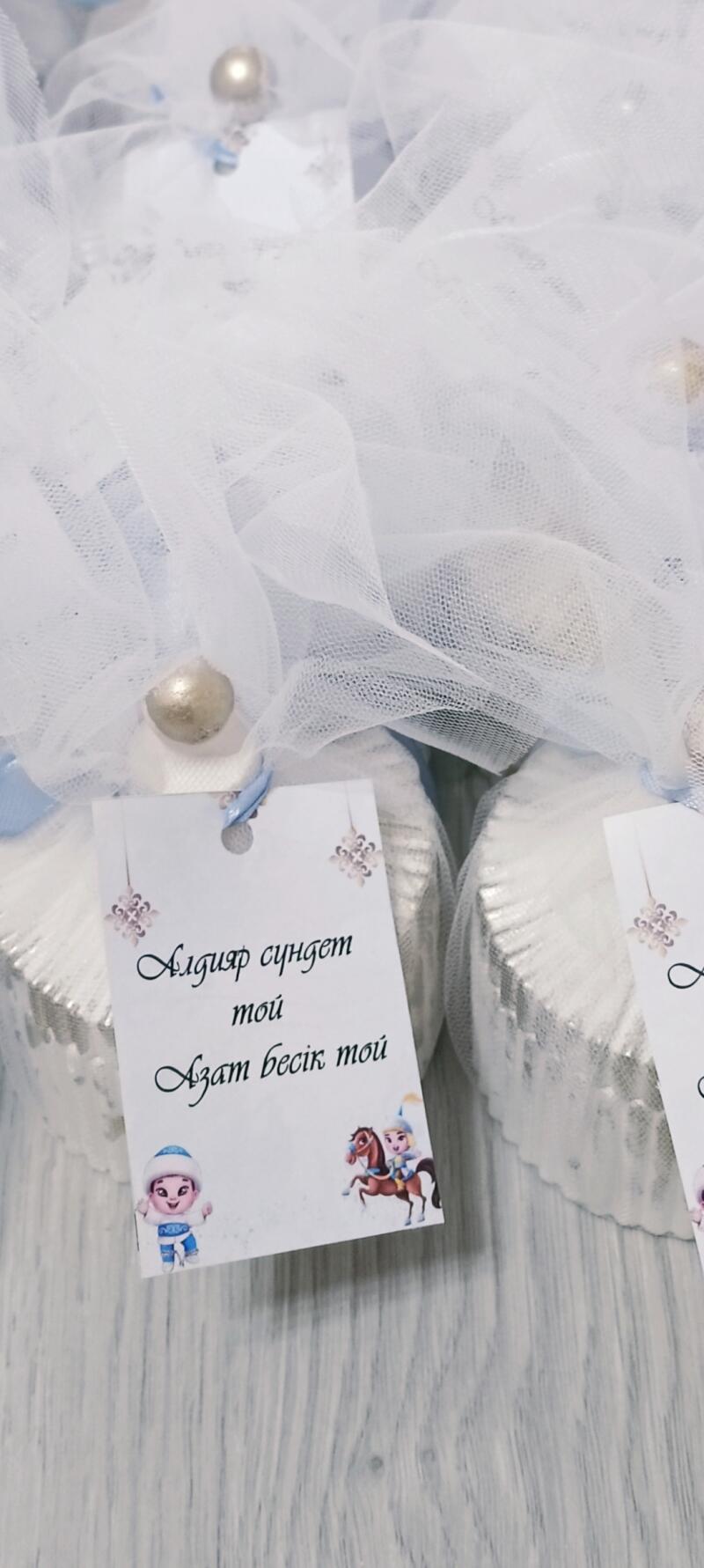The image features small, wrapped favors adorned with white tulle and silver accents. Each favor has a tag that reads "Алдияр сүндет той Азат бесік той" with cartoon-style illustrations.