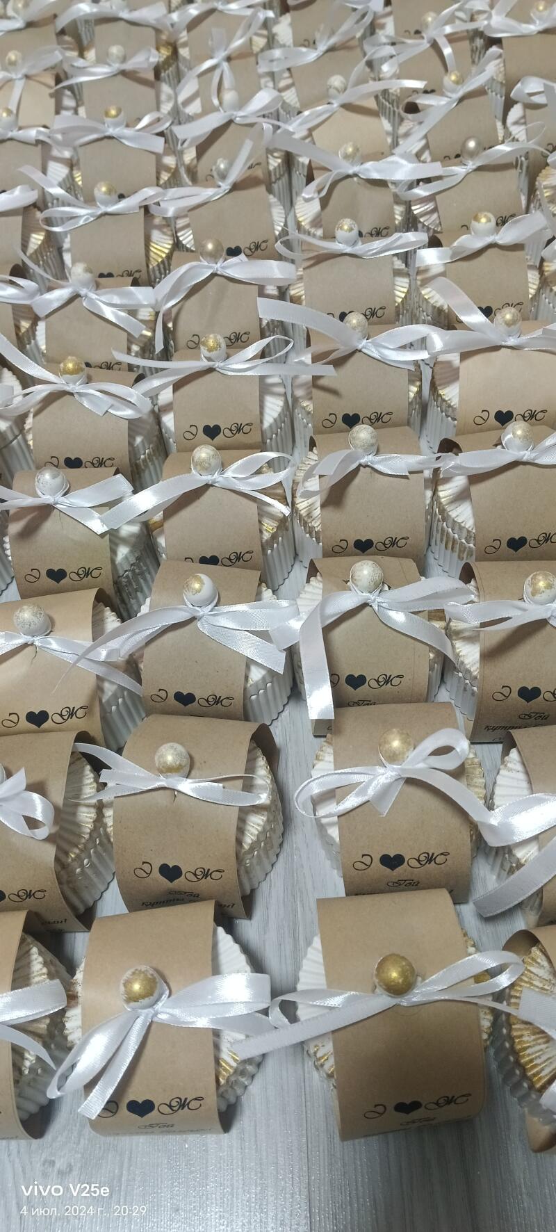 The image shows a large collection of small, decorated gift boxes, each containing what appears to be a cupcake inside a fluted liner, wrapped with a light brown paper band, tied with a white ribbon, and adorned with a small decorative bead. The paper bands have a printed design that includes a heart and the letters "NC".