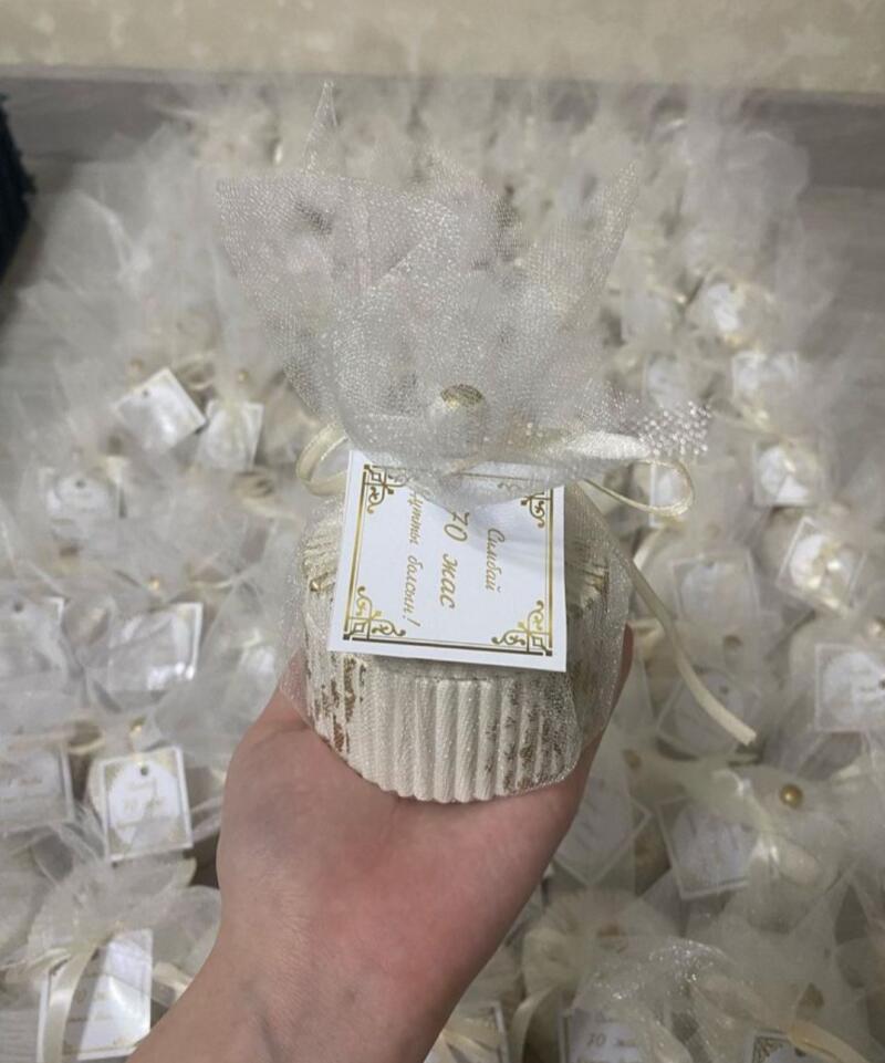 A hand holds a cupcake-shaped favor wrapped in white tulle, adorned with a tag that reads "Симбай 70 жас құтты болсын!", surrounded by many similar favors.