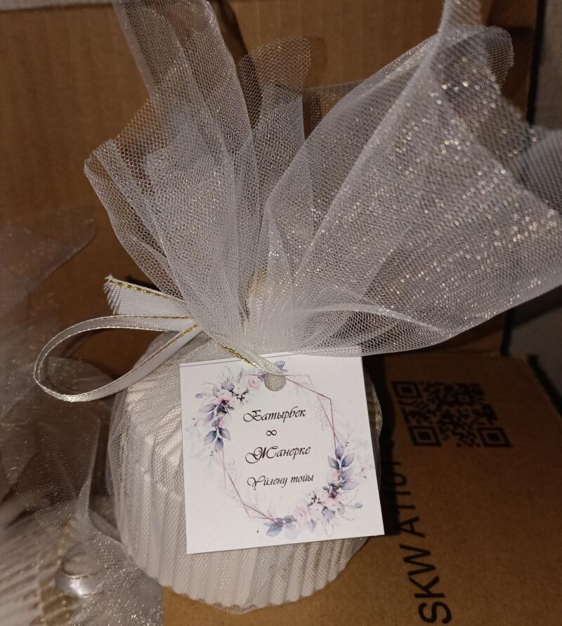 A wedding favor wrapped in white tulle with a ribbon and a tag that reads 'Батырбек и Жанерке, Үйлену тойы' is shown.
