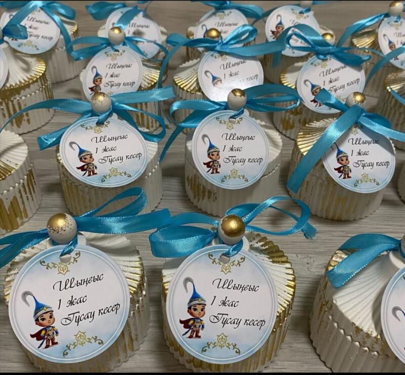 The image shows a collection of small, round decorative boxes, possibly containing treats or favors, each adorned with a turquoise ribbon, a wooden bead, and a tag featuring a cartoon character and the text "Шыңғыс 1 жас Тұсау кесер."