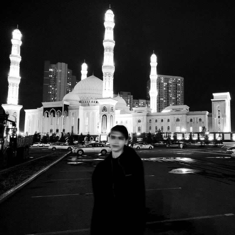 A monochrome shot captures a person standing on an asphalt surface, with a large mosque illuminated in the background. Buildings and parked cars are also visible in the scene.