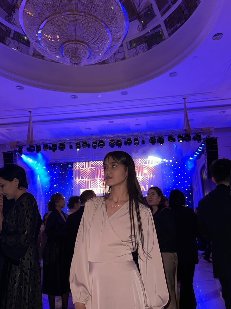 A woman with long dark hair is standing in a crowded room with blue lighting, wearing a light-colored dress; a large chandelier hangs from the ceiling.