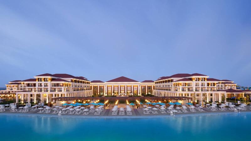 A wide shot shows a large, upscale resort with multiple buildings, red tile roofs, and a long row of white umbrellas and beach chairs along a sandy area that meets a large pool of blue water under a clear sky.