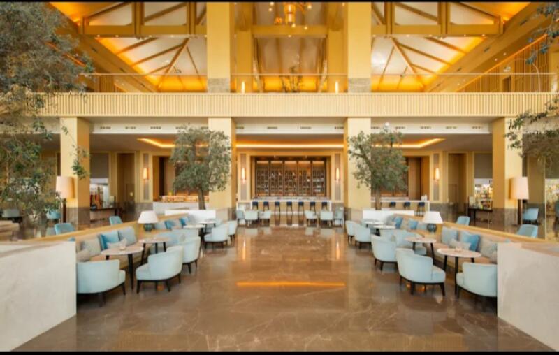 The photo shows the interior of a large lobby with light blue seating areas on both sides, trees, and a bar in the background.