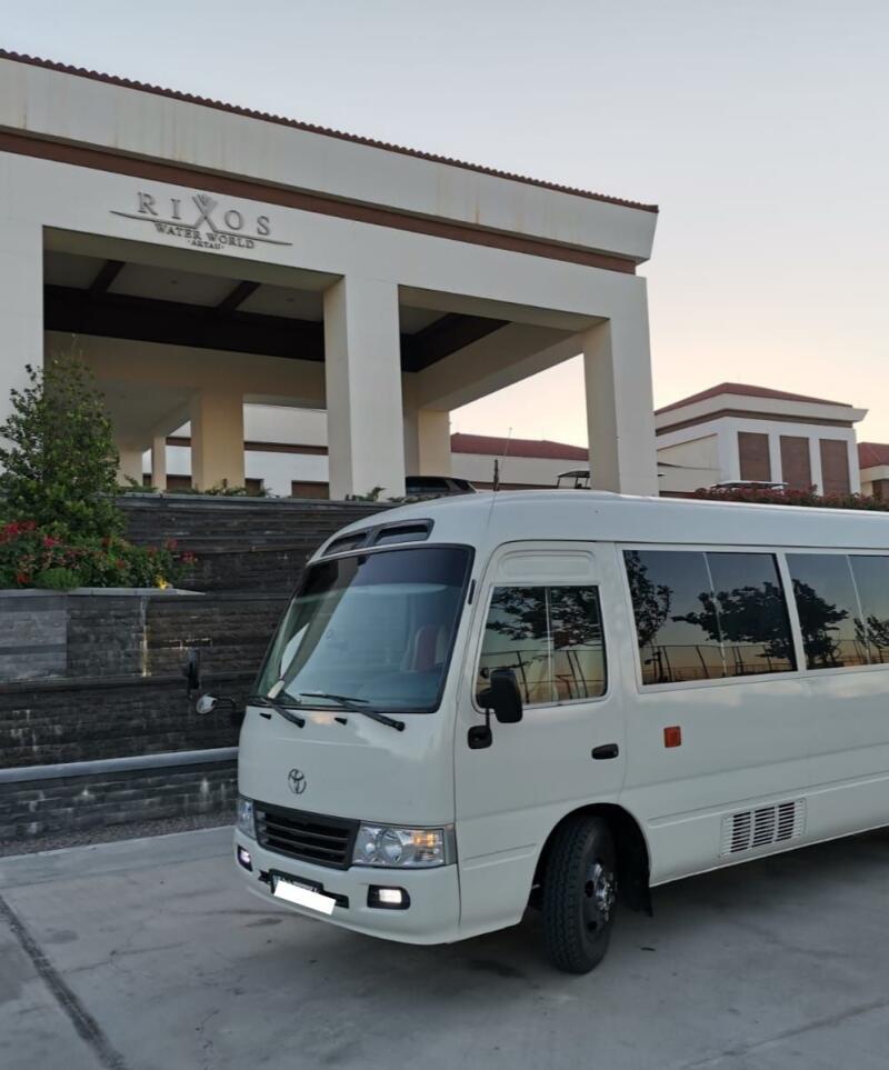 A white Toyota Coaster bus is parked in front of the Rixos Water World hotel building.