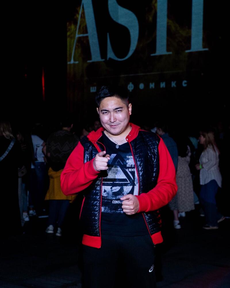 A man in a red and black jacket points at the camera with a crowd of people and the word "ASTI" in the background.