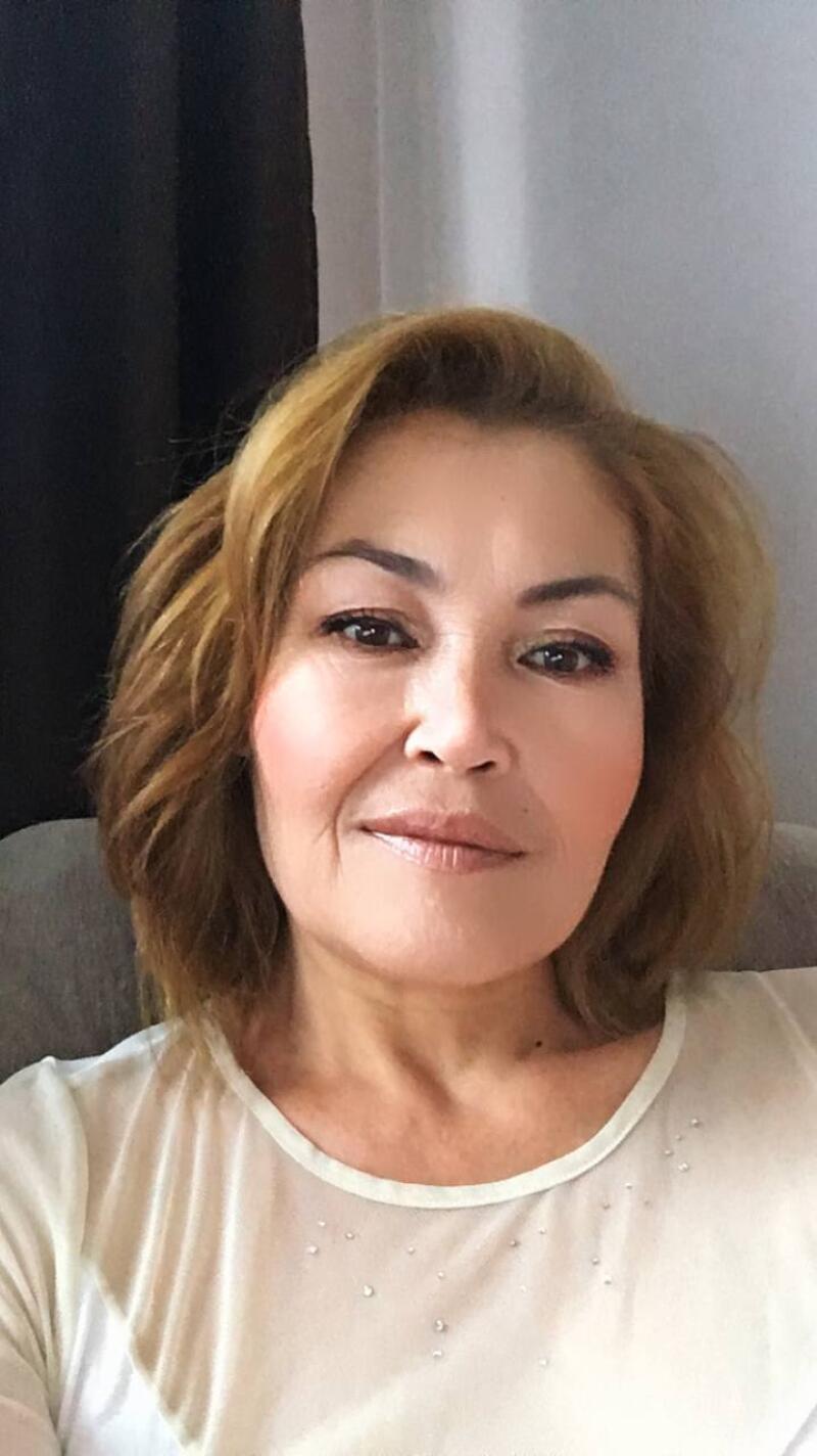 A woman with shoulder-length brown hair and fair skin is wearing a sheer white top with small embellishments. She has makeup on and is looking directly at the camera.