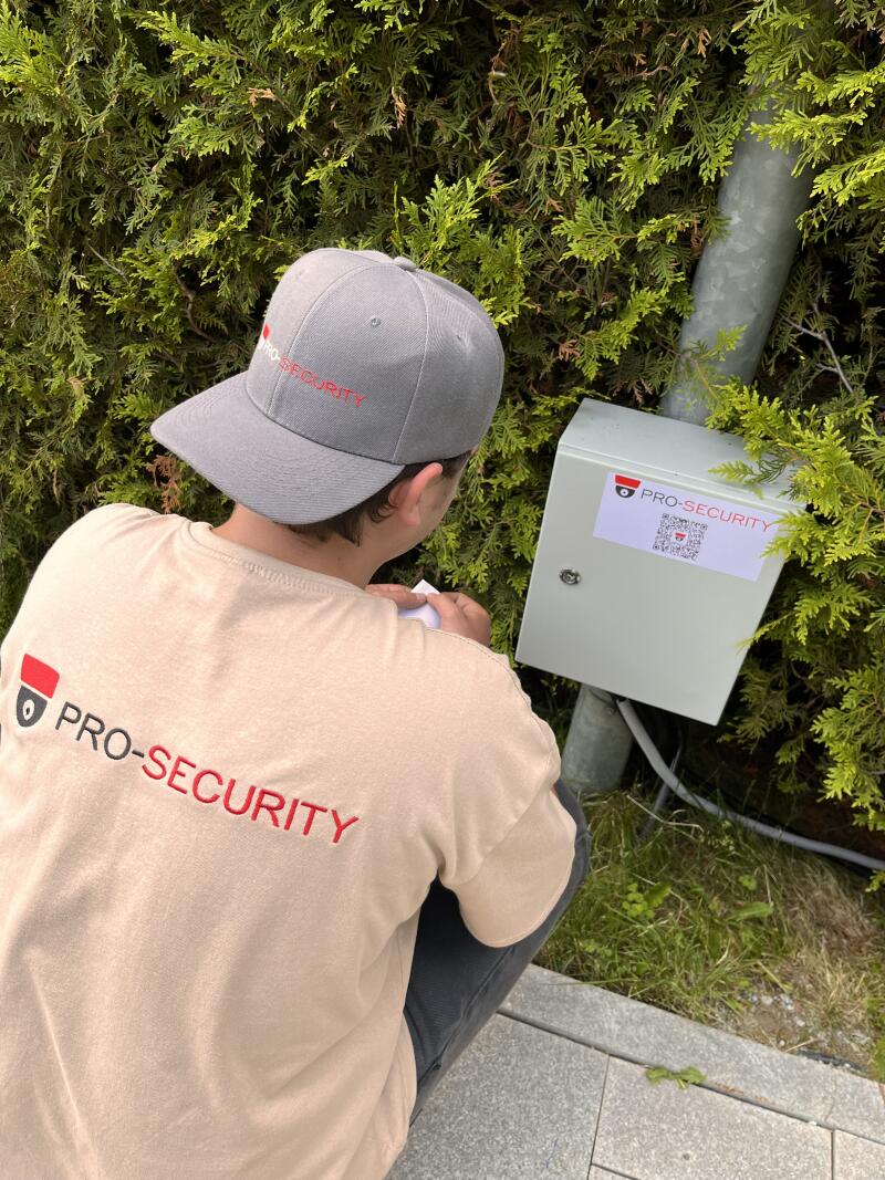 A person wearing a Pro-Security hat and shirt squats next to a gray box with a Pro-Security sticker and QR code on it, set against a backdrop of green bushes.