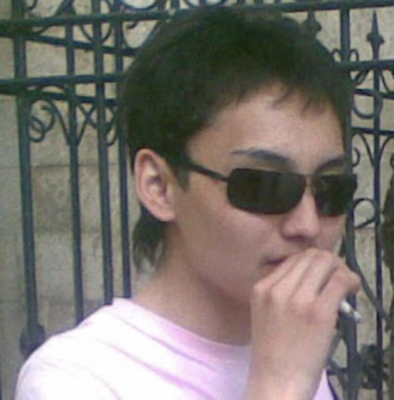 A person with dark hair and sunglasses holds a cigarette in their hand, standing in front of an ornate metal fence.