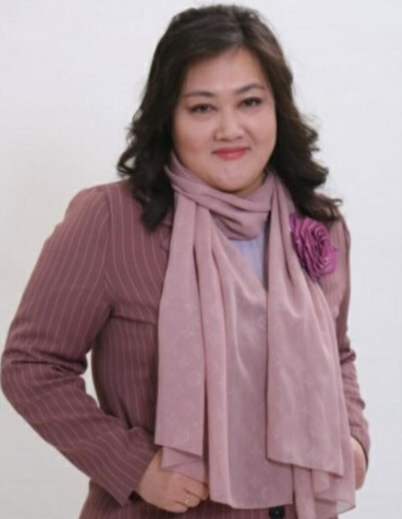 A woman with dark wavy hair is wearing a striped pink suit with a matching scarf and a large rose pin.