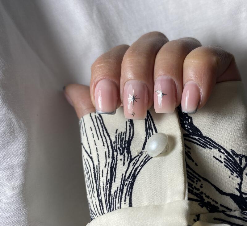 A hand with manicured nails is seen against a sleeve with a button; the nails are nude with silver tips and star decals.