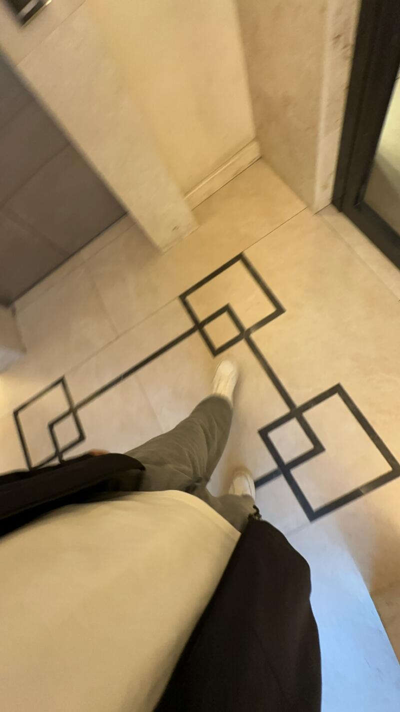 A person wearing white sneakers and grey pants is standing on a tile floor with a geometric black pattern, also wearing a white shirt and black jacket.