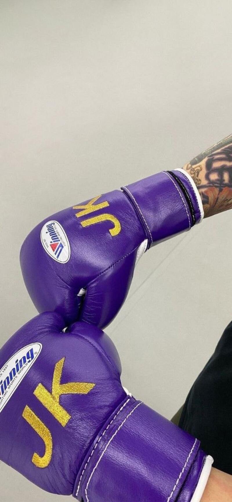 A close-up shows two purple Winning boxing gloves with "JK" stitched in gold. The gloves are stacked, one on top of the other, and the arm of the person wearing the gloves is visible, showing tattoos.