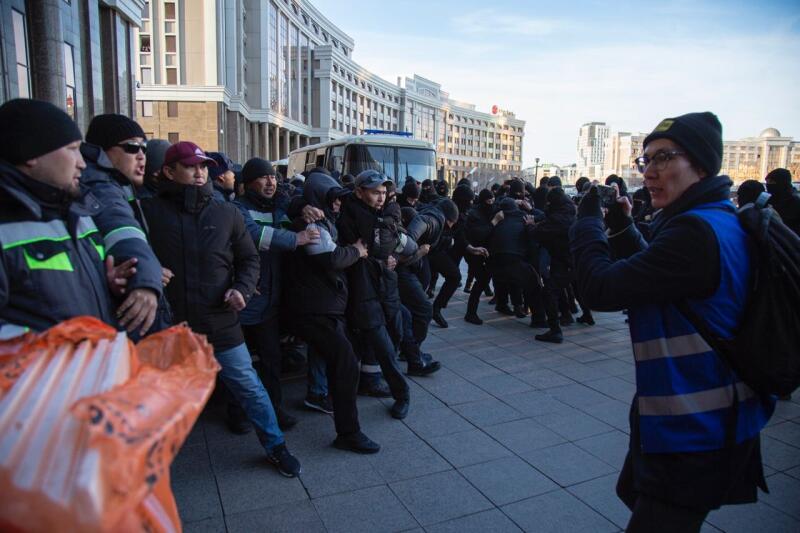 A crowd of people are being apprehended by police officers in riot gear, while a woman in a blue vest and glasses takes photos of the event.