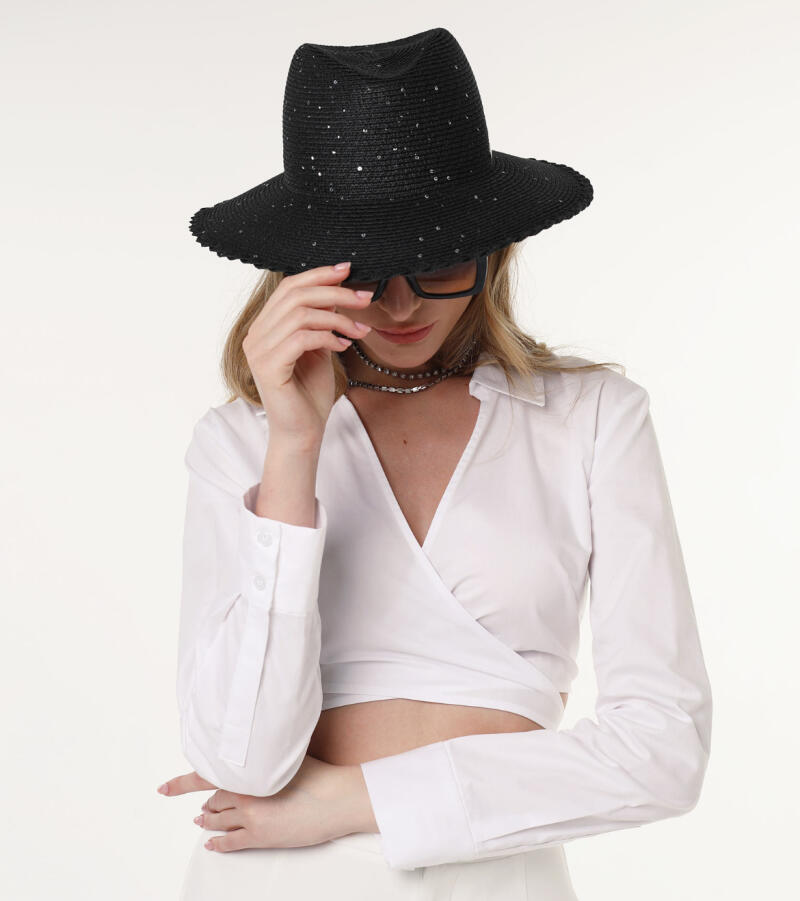 A woman in a white crop top, sunglasses, and a black, sparkly hat is posing with her head down.