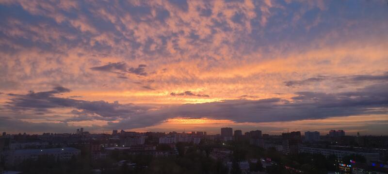 The photo captures a cityscape silhouetted against a vibrant sunset sky with clouds colored in shades of orange, pink, and blue.