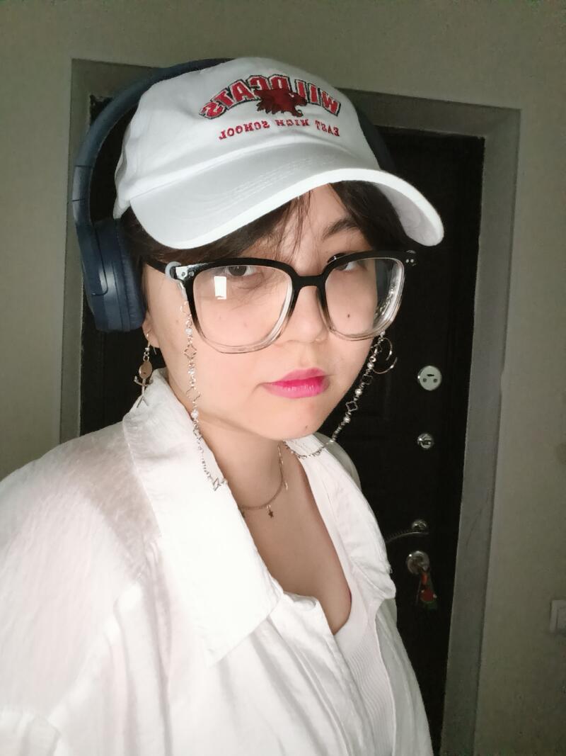 A person wearing glasses, headphones, and a white cap with the text "WILDCATS HIGH SCHOOL" is seen in a close-up shot; they are also wearing a white shirt and a necklace.