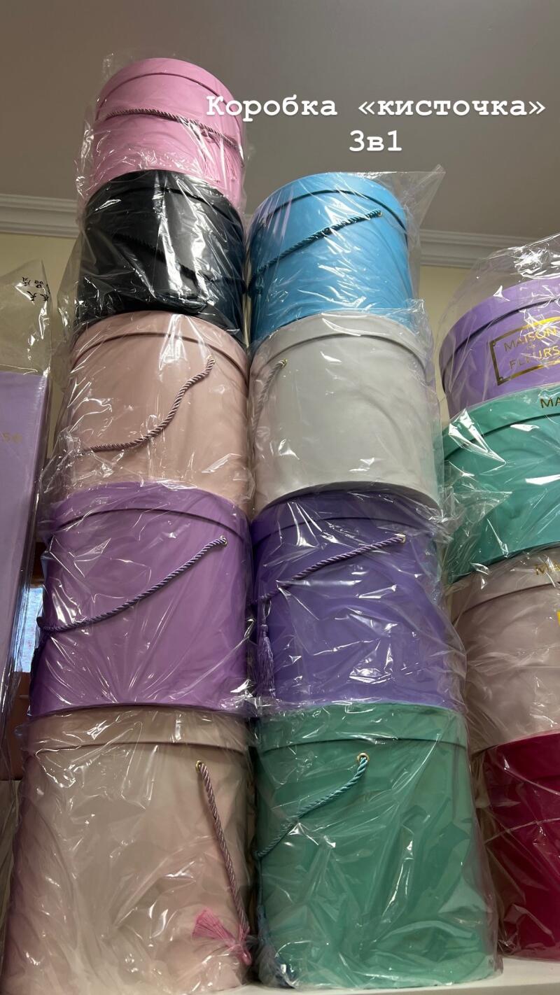 A stack of round gift boxes in various pastel colors wrapped in clear plastic, with visible text that reads "Коробка «кисточка» 3в1."