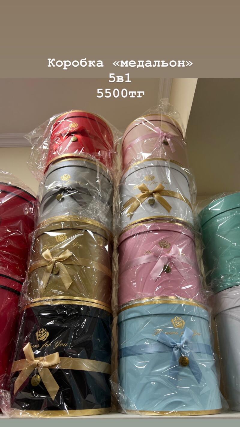 The image shows a stack of round gift boxes in various colors, each wrapped in plastic and adorned with a ribbon and a small tag. Text visible includes "Коробка «медальон»", "5в1", and "5500тг".