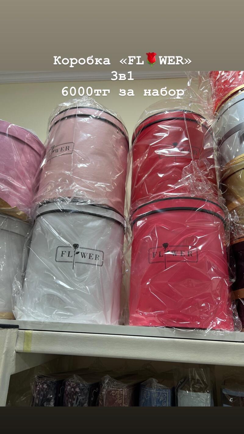 The image shows stacked round boxes of different colors wrapped in cellophane; the text on the boxes reads "FLOWER" with a floral logo, and the text above them reads "Коробка FLOWER 3в1 6000тг за набор".