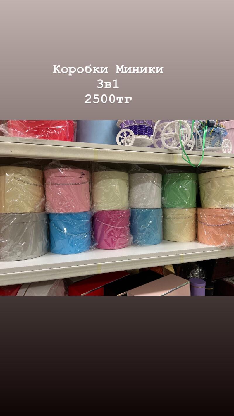 The image shows shelves filled with colorful, round gift boxes wrapped in clear plastic, with the text "Коробки Миники 3в1 2500тг" displayed above.