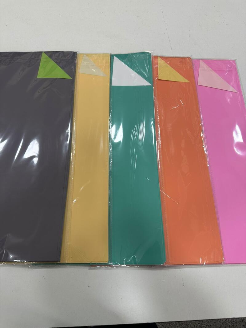The image shows a row of colorful paper sheets in plastic packaging, each sheet topped with a triangular piece of colored tape.