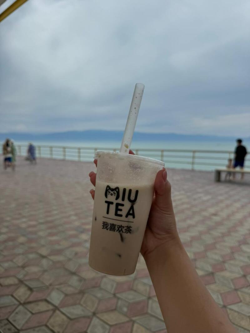 A hand holds a plastic cup with a straw filled with milk tea from "MIU TEA" against the backdrop of the sea and cloudy sky; the cup has a logo and the text "我喜欢茶".