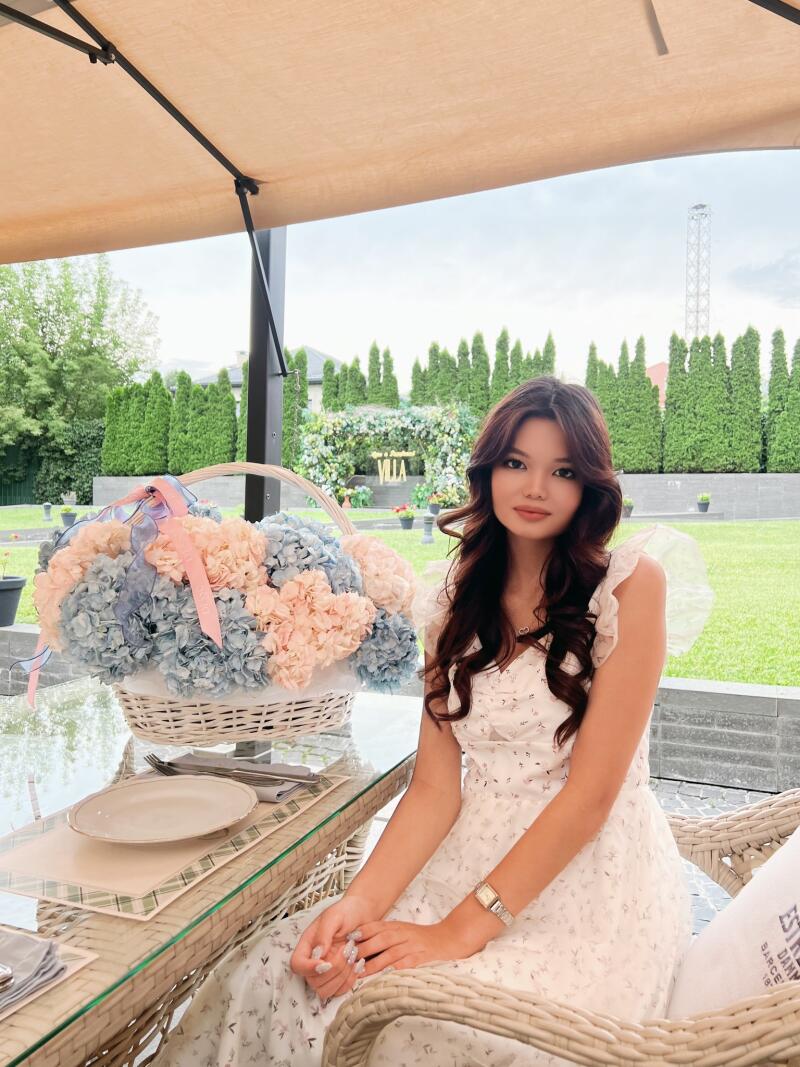 A young woman in a white floral dress sits at a glass table, next to a white basket full of pink and blue flowers.