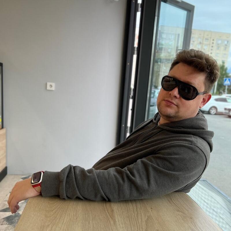 A man with sunglasses and a hoodie is leaning on a table, wearing a red watch.