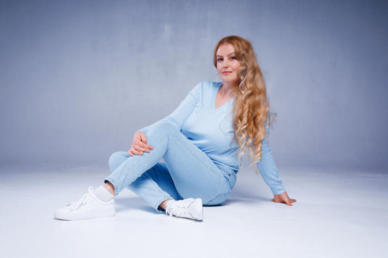 A woman with long, wavy blonde hair is sitting on the floor wearing a light blue V-neck shirt, light blue jeans, and white sneakers.