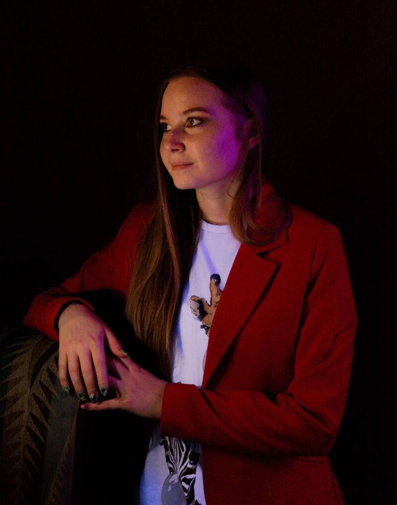 A woman with long hair in a red blazer poses in front of a dark background, wearing a white t-shirt with a giraffe and zebra print.