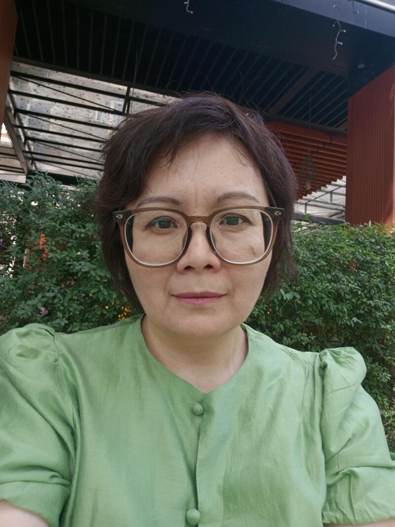 A woman with short dark hair and glasses is wearing a green button-up shirt. She is standing outside with greenery visible in the background.