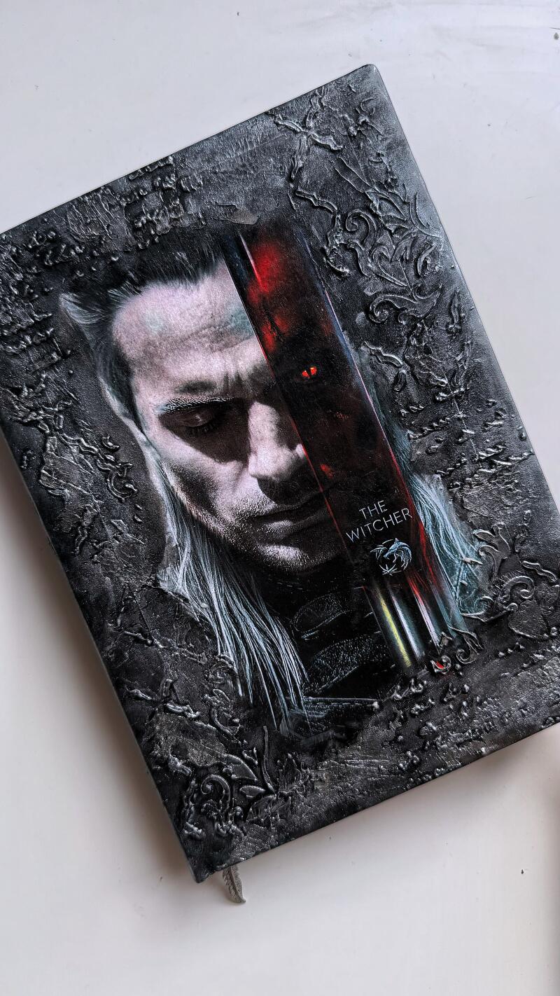 A close-up shows a textured, dark-colored book cover featuring an image of Geralt of Rivia from "The Witcher."