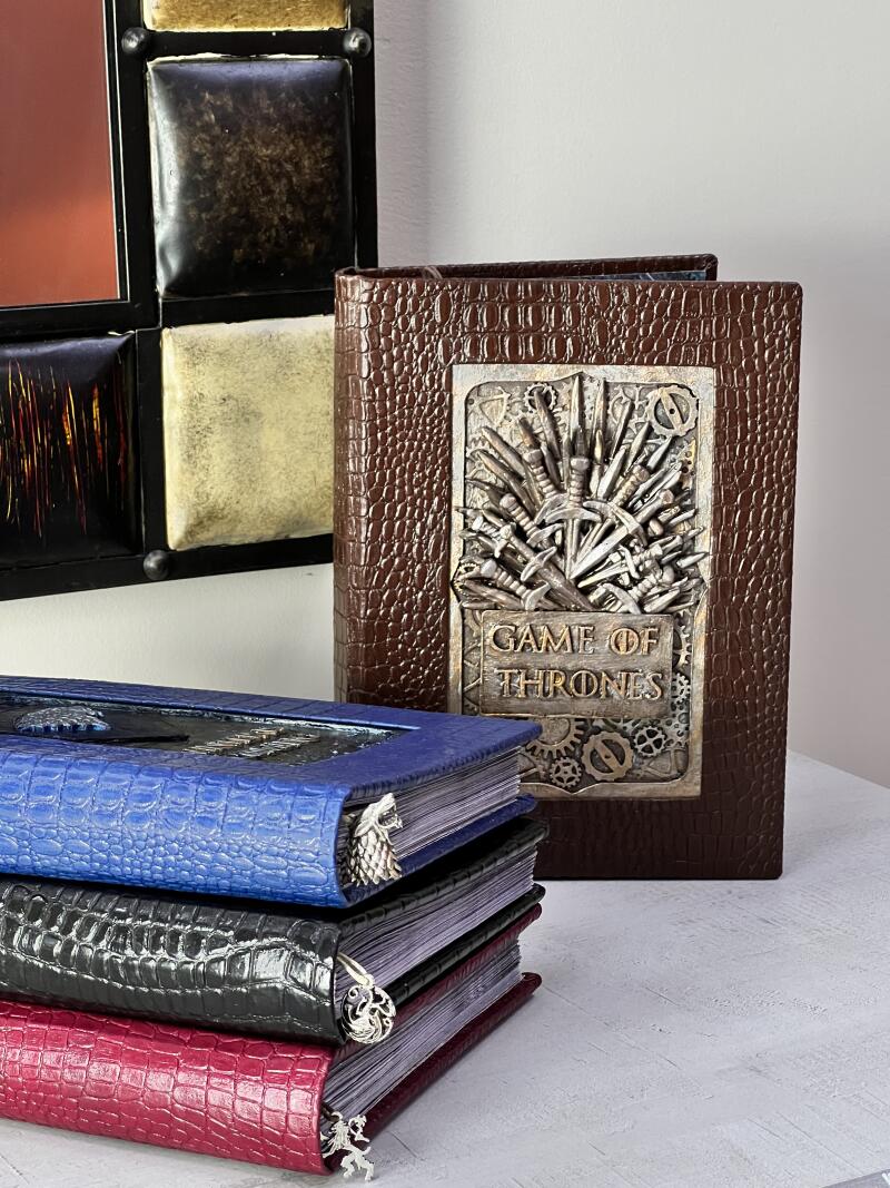 The image shows a stack of three books with a brown "Game of Thrones" book behind them. The books are blue, black, and red, all with a reptile skin texture and a metal charm bookmark. The "Game of Thrones" book is brown with a metal emblem of swords and gears with the title "Game of Thrones" displayed.