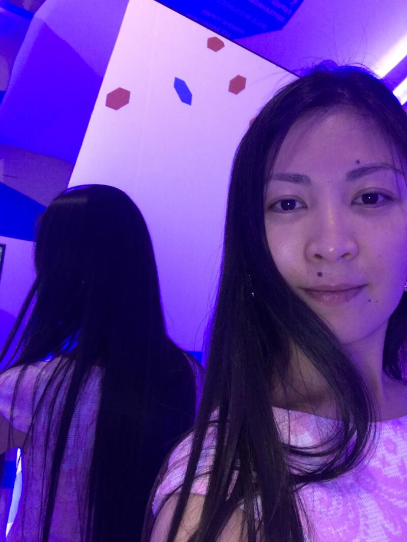 A woman with long dark hair stands in front of a mirror under blue and purple lighting, wearing a pink patterned shirt.