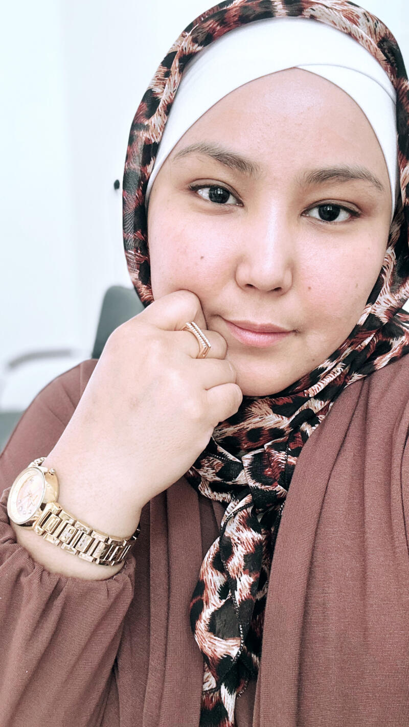 A woman in a hijab poses with a gold watch and ring, wearing a brown dress and a scarf with an animal print.