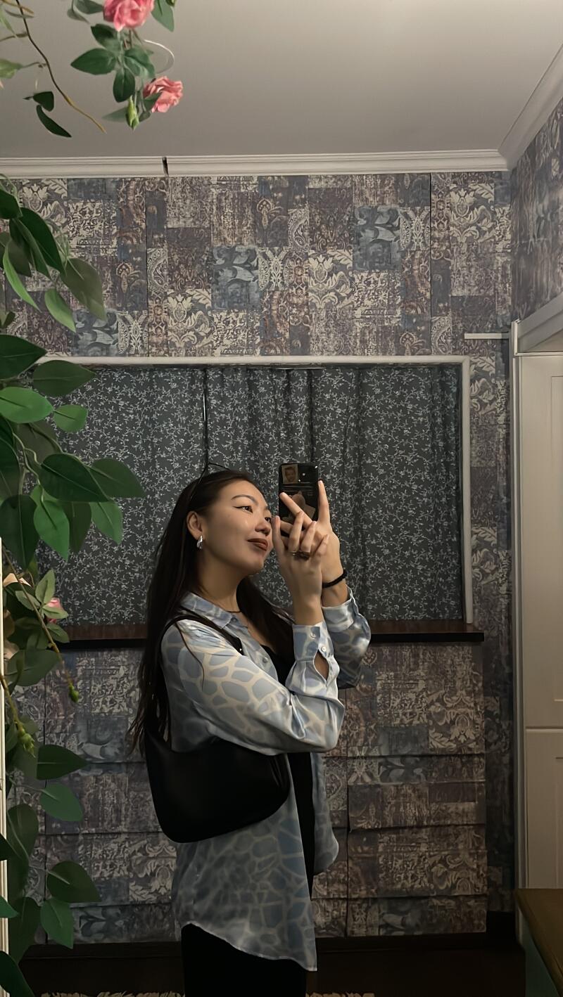 A woman with long dark hair and a light blue patterned blouse takes a selfie in a mirror, with a black purse slung over her shoulder.