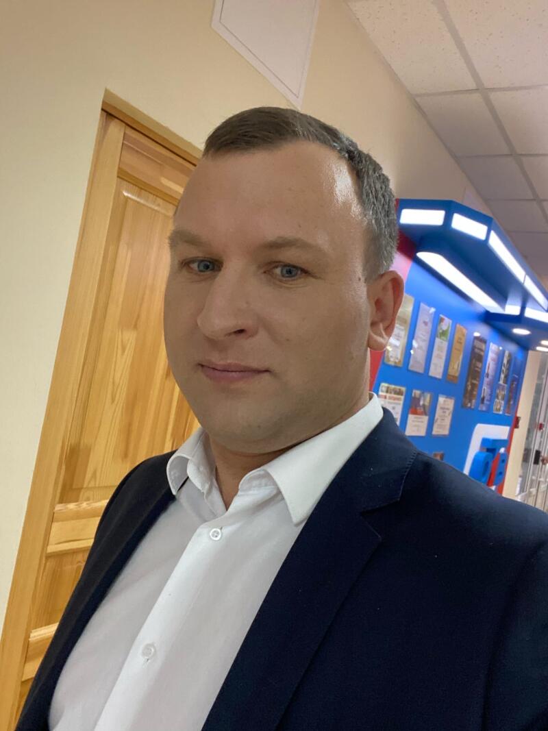 A man with short hair and blue eyes is wearing a white shirt and a navy blue suit jacket. He is in a hallway with a wooden door to his left and a blue display case behind him.