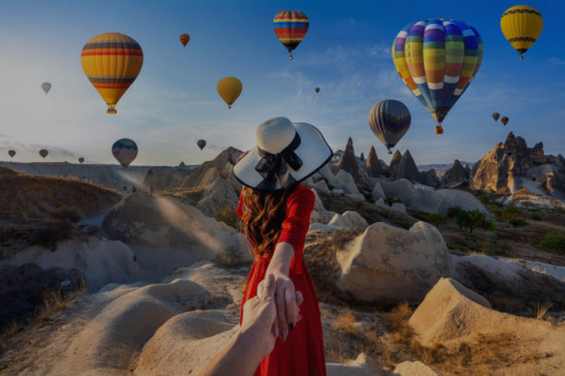 A woman in a red dress and wide-brimmed hat leads someone by the hand through a landscape dotted with rock formations, with several hot air balloons floating in the sky.
