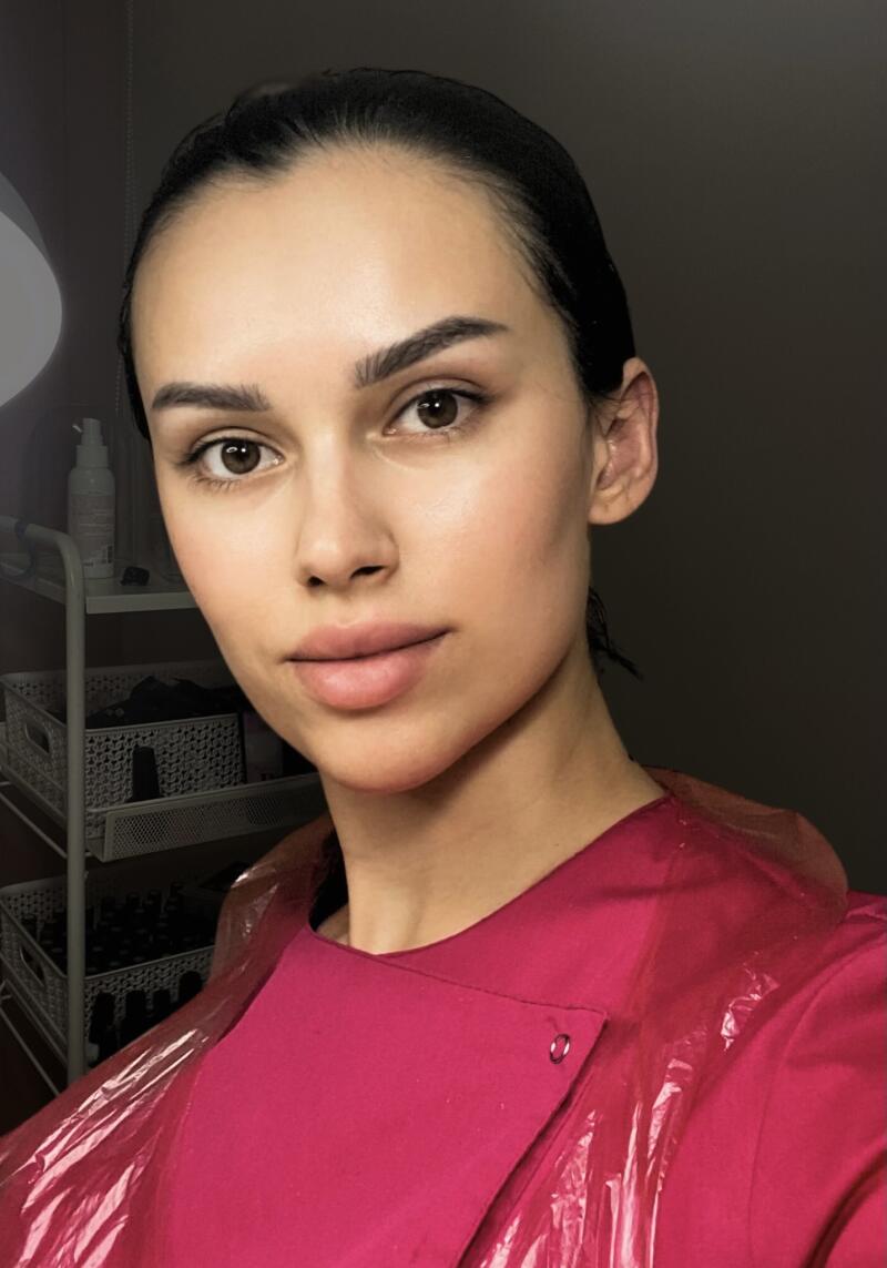 A woman with dark hair and brown eyes poses wearing a fuchsia top with a plastic covering, with makeup applied and a shelf with baskets visible in the background.