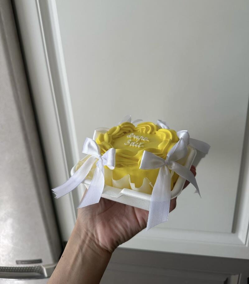 A hand holds a small, yellow cake with white icing that reads 'Love Salt', adorned with white ribbons, and packaged in a white container.
