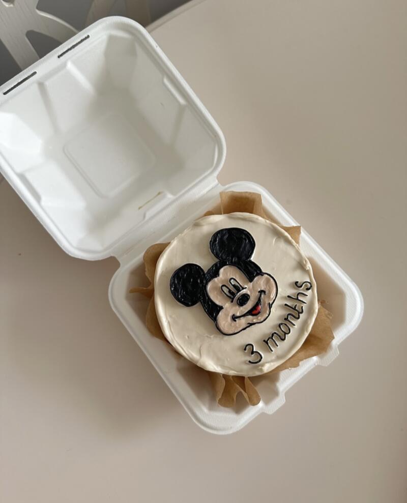 A small cake with white frosting and a Mickey Mouse design sits inside a white takeout container, with the words '3 months' written on the cake.