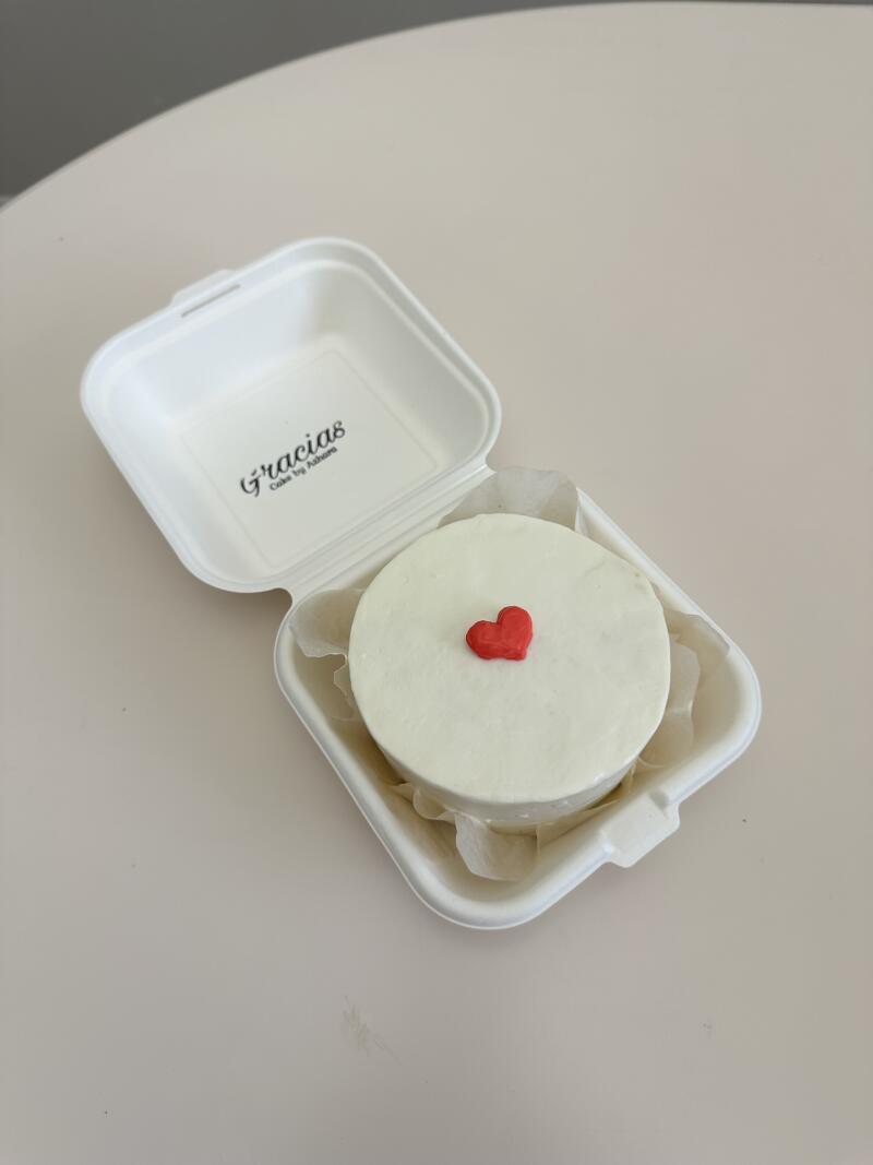 A small, round, white cake with a red heart on top is nestled in a white takeout container lined with parchment paper, with the text "Gracias Cake by Adhore" visible inside the container.