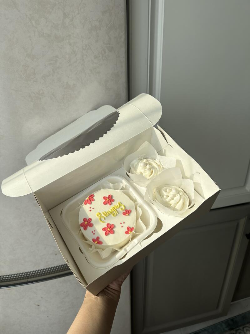 A white box contains a small cake decorated with pink flowers and yellow lettering that says "8 наурыз," along with two cupcake-sized portions of white frosting.