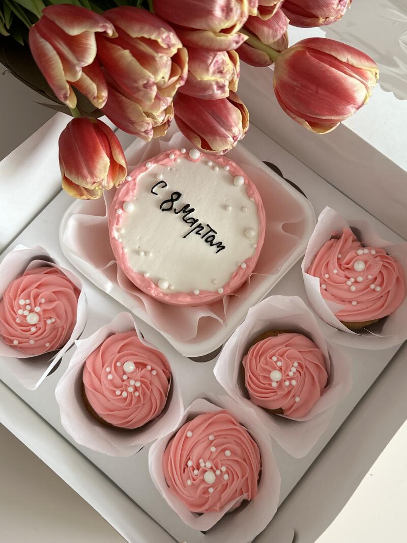 A box contains a small cake with the inscription "С 8 Марта" (Happy March 8th) and several cupcakes with pink frosting, decorated with white sprinkles; pink tulips are arranged around the edge of the box.