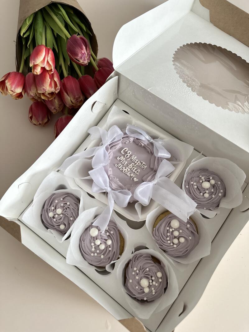 A box contains a cake with the inscription "March 8 We love you" and cupcakes with lilac cream and white sprinkles, with a bouquet of tulips nearby.