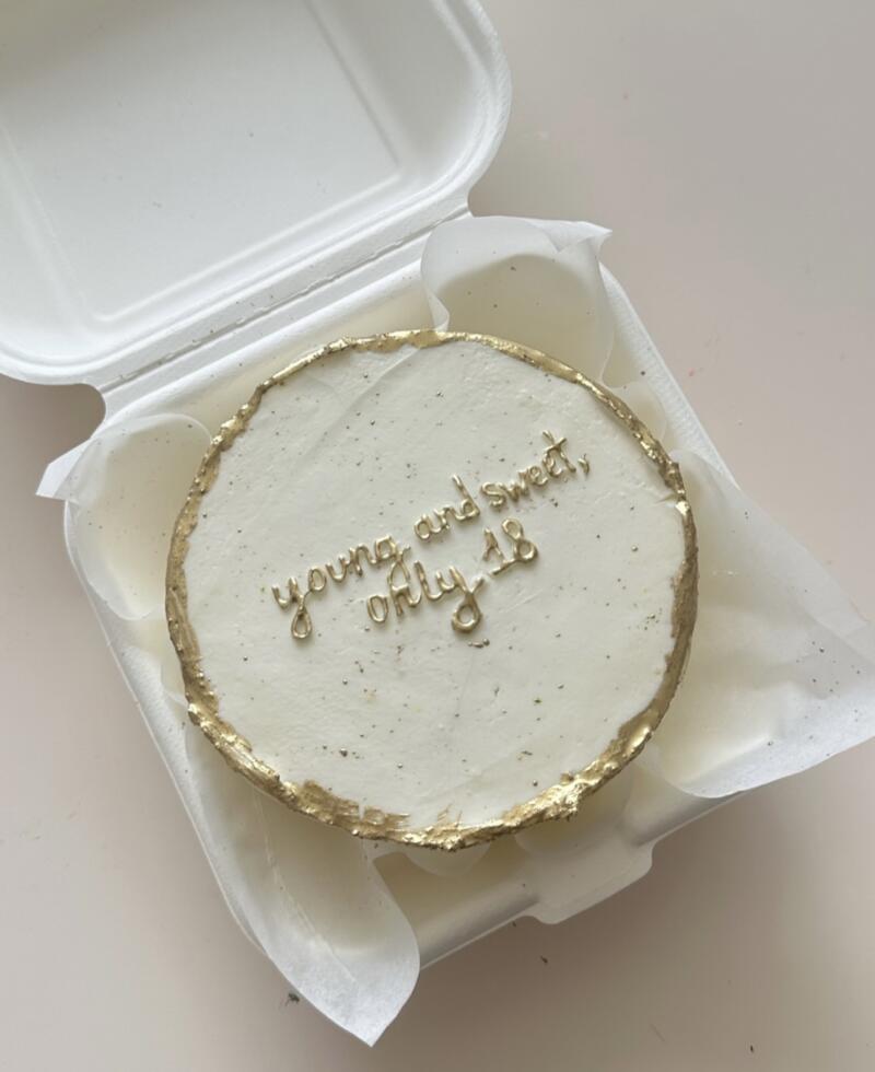 A small, round cake with white frosting and a gold leaf border sits inside an open white takeout container; the cake is inscribed with gold cursive that reads, "young and sweet, only 18".