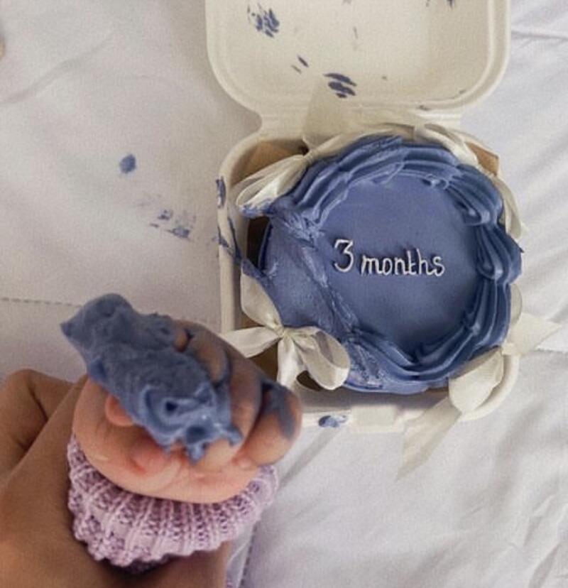 A hand holds a small, blue-iced figure over a pink knitted cuff, with a small cake decorated with blue icing and the words "3 months" visible in the background.