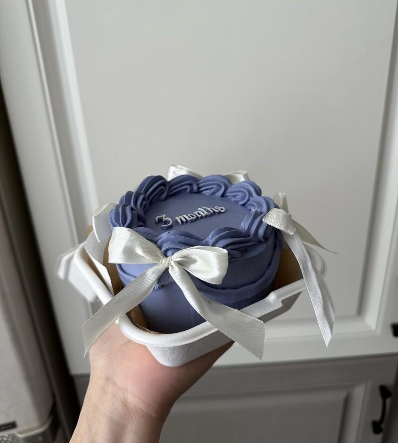A hand holds a small, lavender-colored cake decorated with white frosting and a white ribbon, with the text "3 months" written on top.